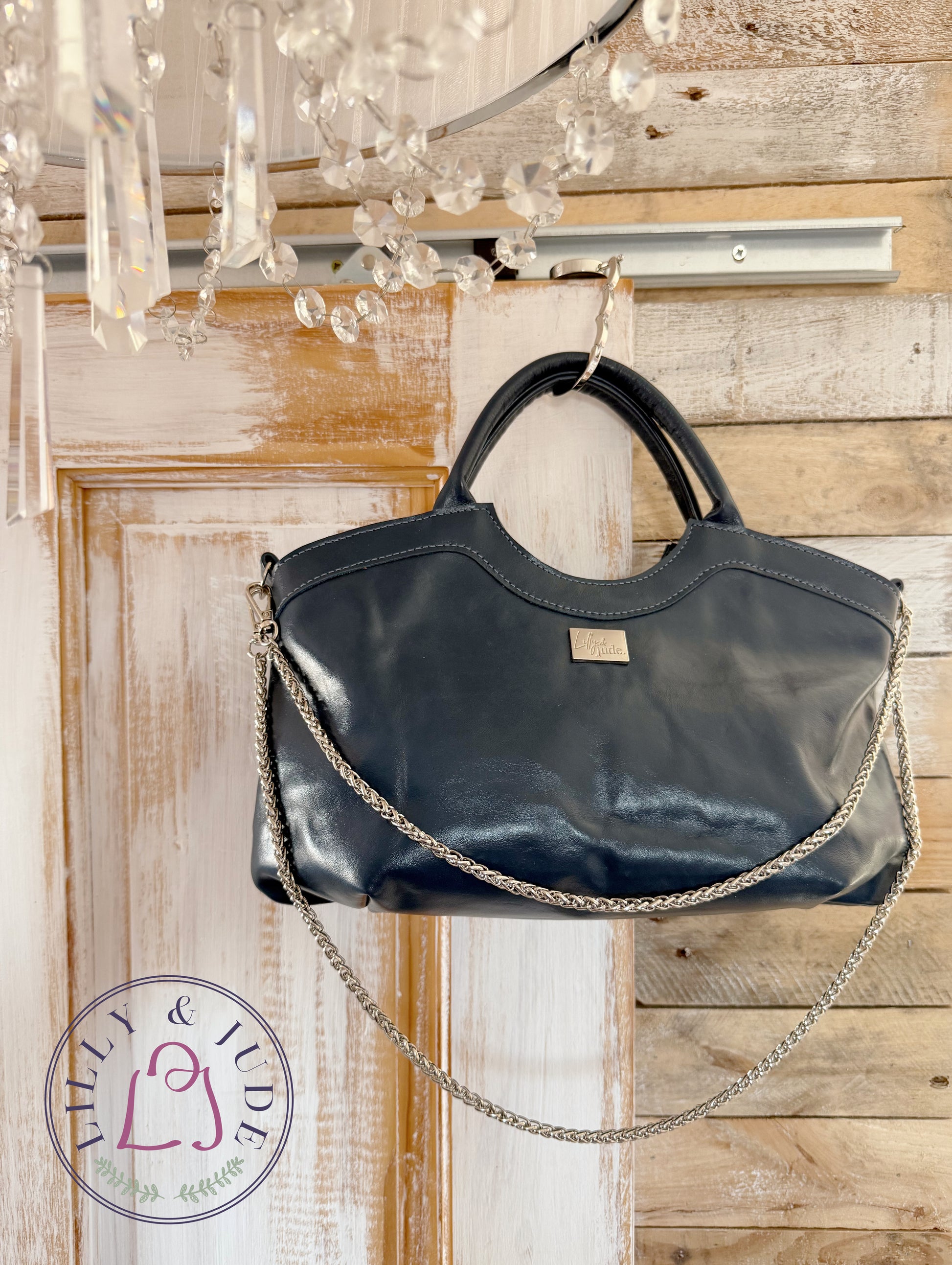 Navy, rescued leather handbag with a chain detail hanging on a wooden door with a chandelier in the foreground.