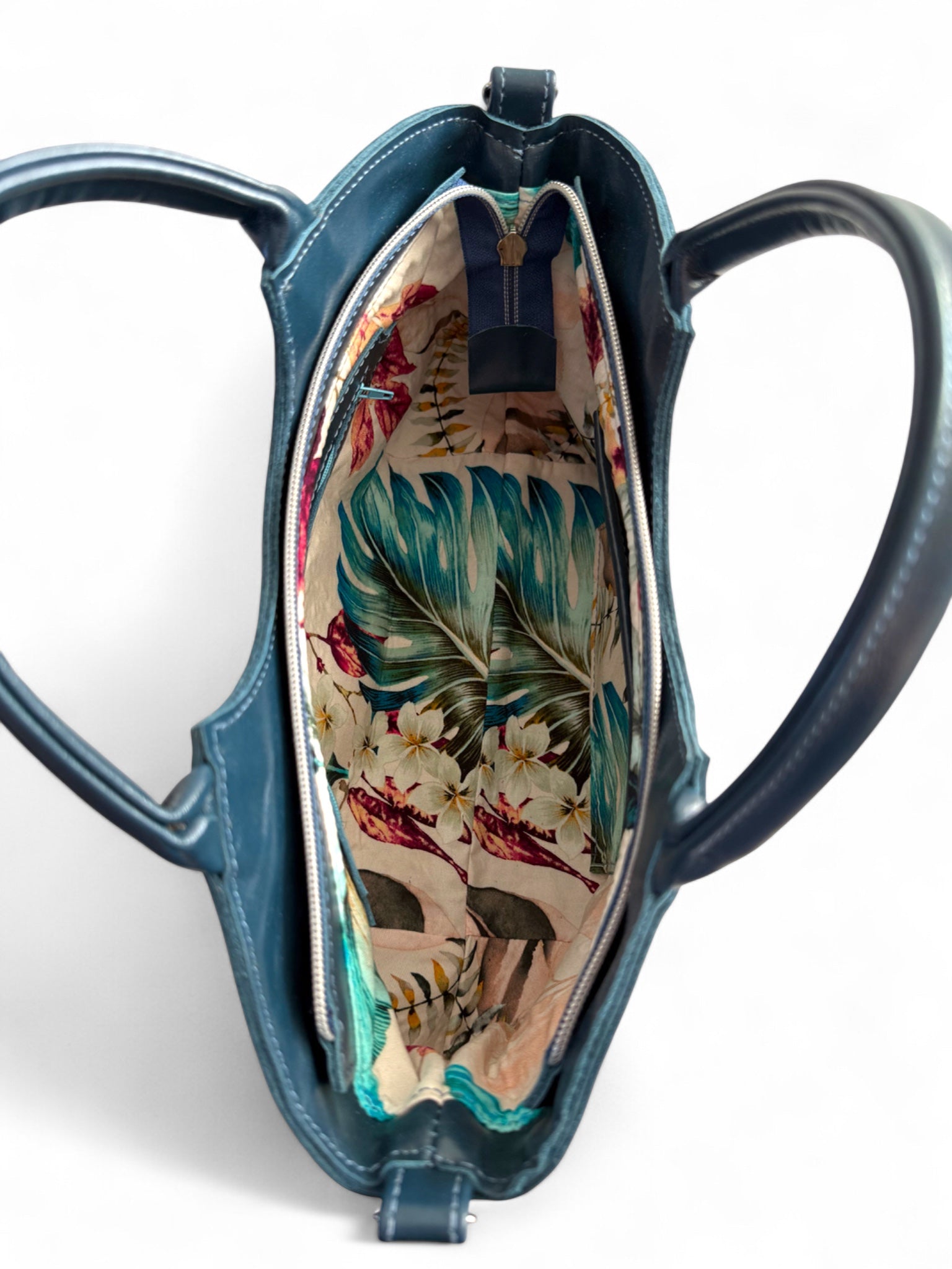 Above view of the navy blue/ dark teal Flossy handbag. The bag is open and is showing the tropical style floral lining fabric inside. The back is sat on a white background