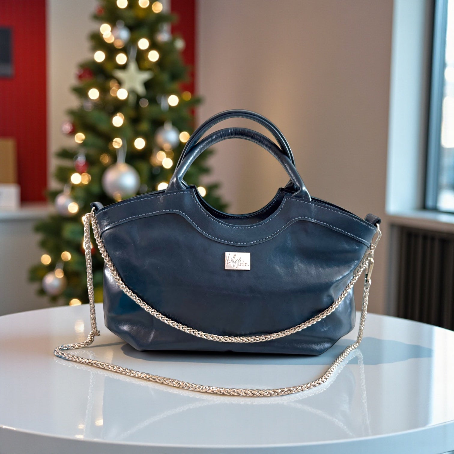 Handcrafted blue leather handbag with a silver chain detail on a table in front of a decorated Christmas tree. Exclusive to Lilly & Jude