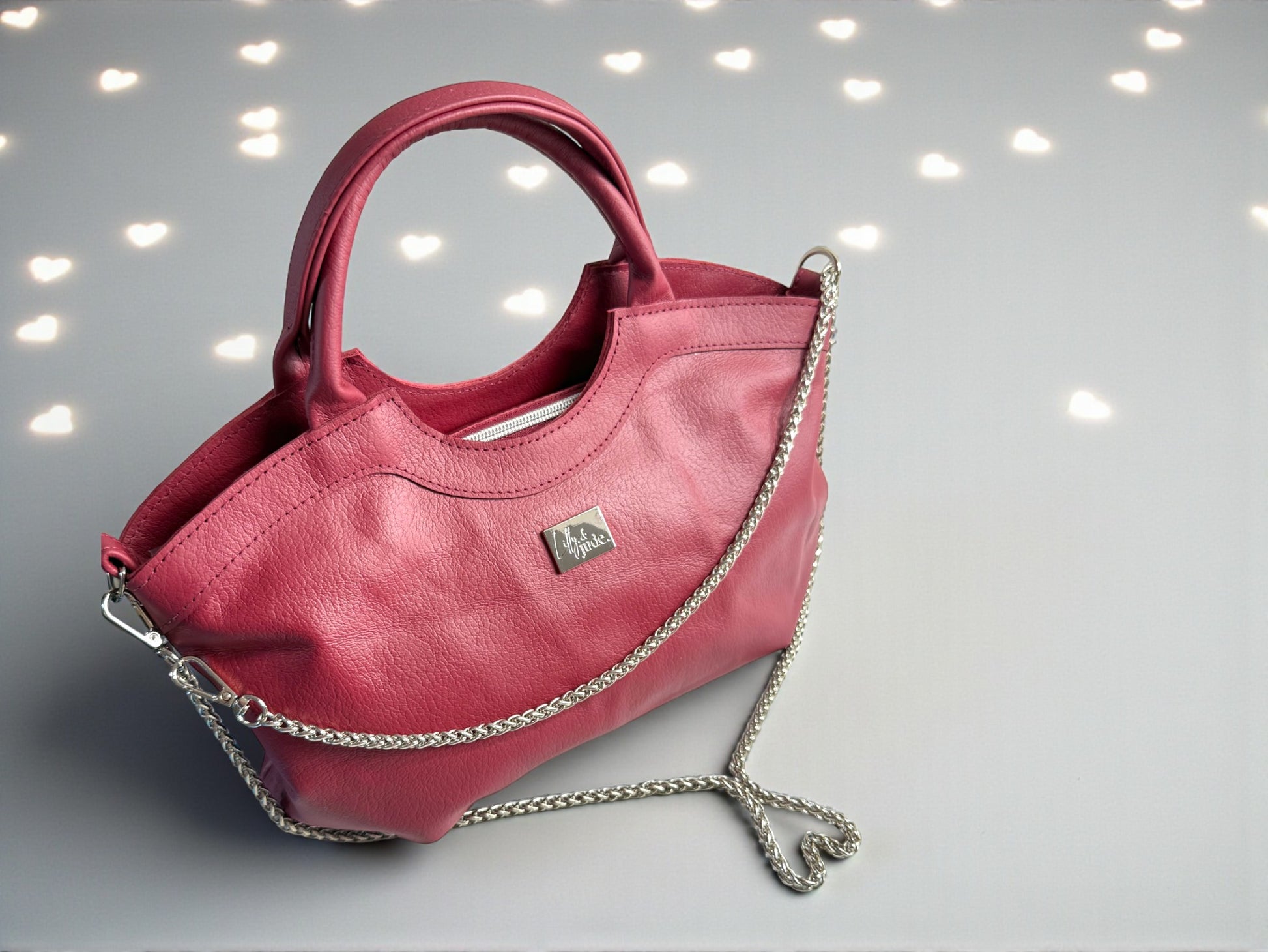 Red leather Flossy handbag with silver chain strap, which is arranged in a heart shape, sitting on a pale grey background, with heart shaped bokeh lighting
