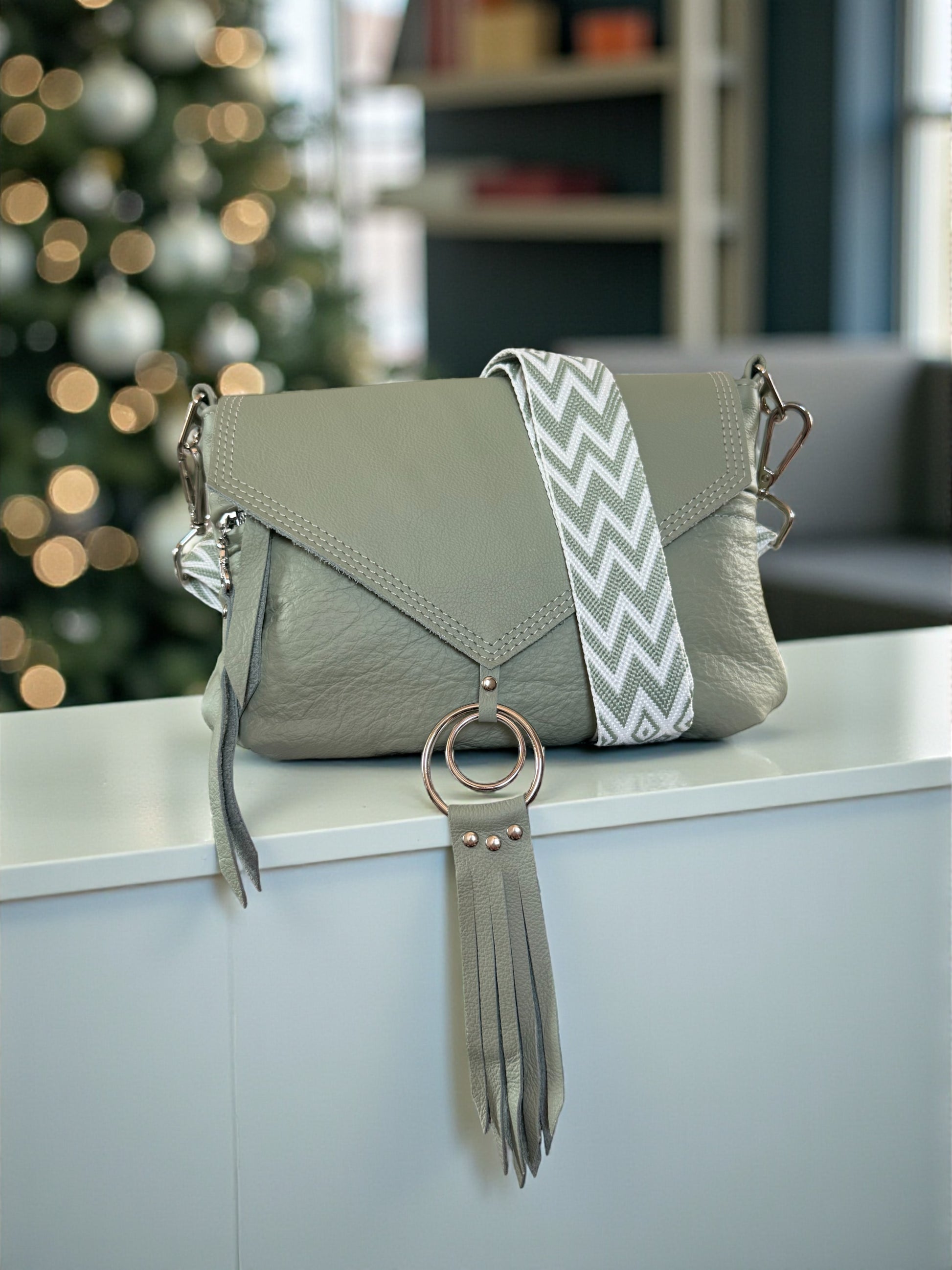 Light green grey  handbag with a decorative strap and tassel on a surface with a blurred festive background