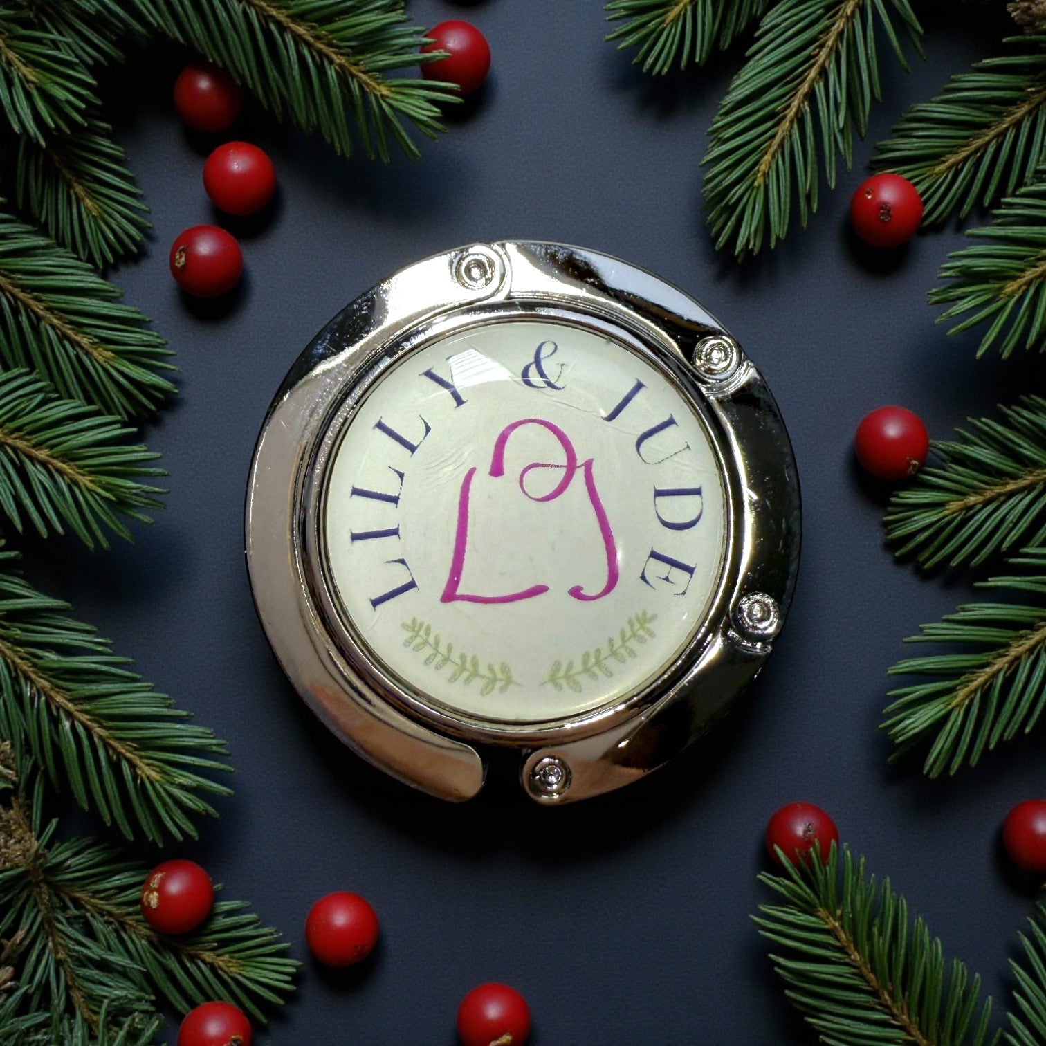Round bag table hook with 'LILLY & JUDE' branding surrounded by pine branches and berries on a dark background
