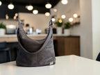 A dark gray leather shoulder bag sits on a white table in a softly blurred cafe or office interior with hanging lights and plants in the background.