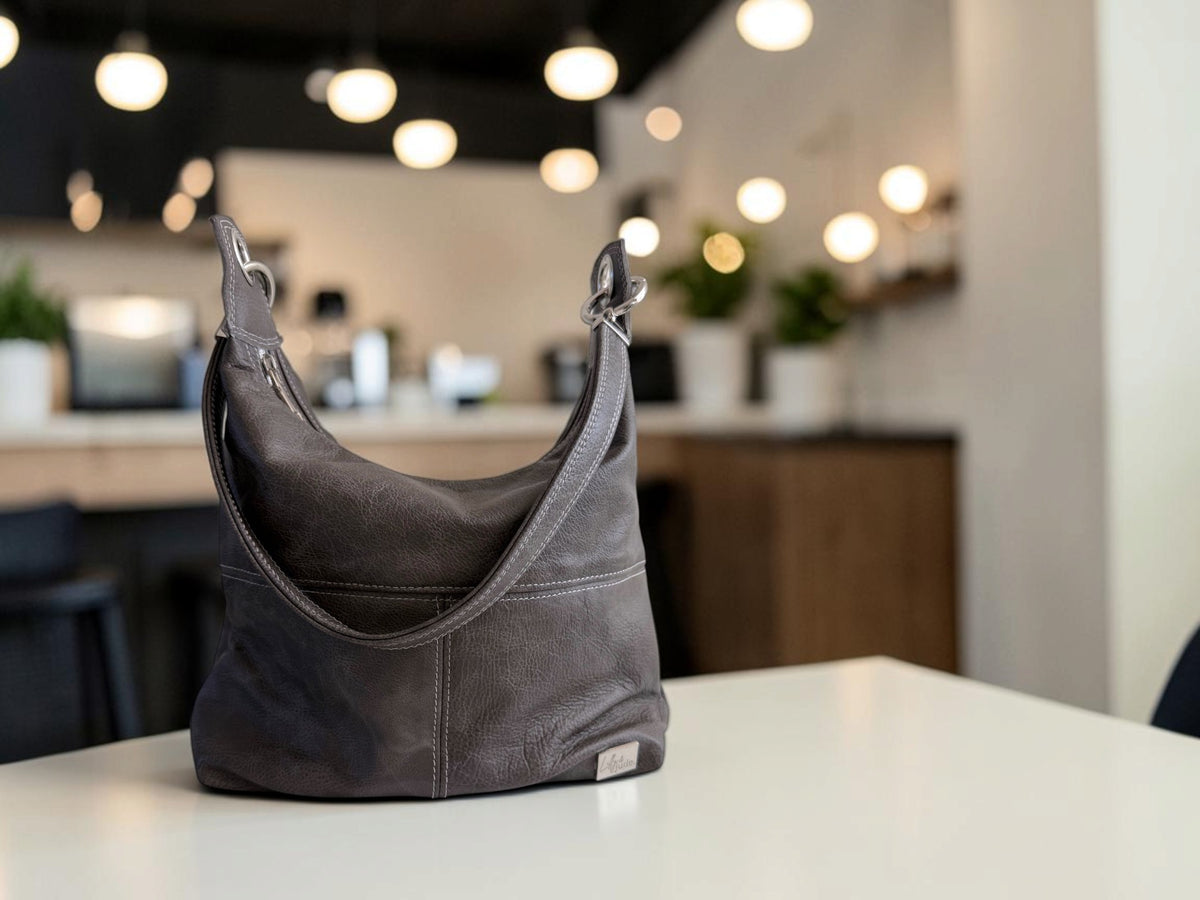 A dark gray leather shoulder bag sits on a white table in a softly blurred cafe or office interior with hanging lights and plants in the background.
