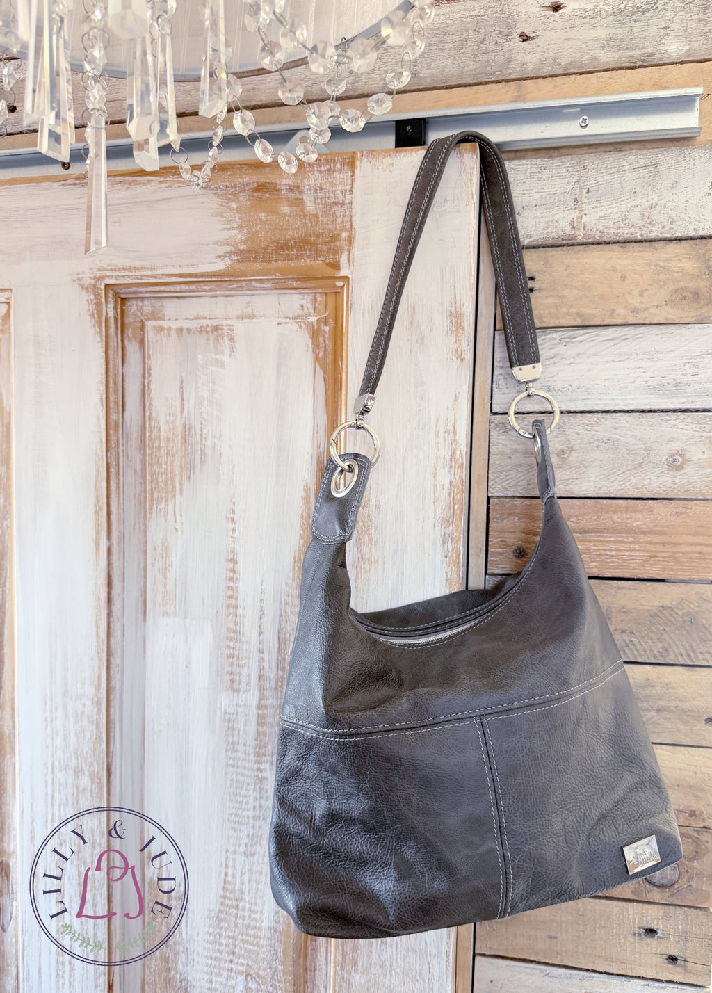 A dark gray leather hobo shoulder bag hanging from a wooden door and wood panel wall. A small brand tag is visible on the lower right corner of the bag.