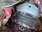 Close-up of the interior of a handbag showing a patterned fabric lining with purple, pink, and beige swirls, a light gray suede flap pocket with a snap button, and a dark grey leather card pocket with white contrast stitching.