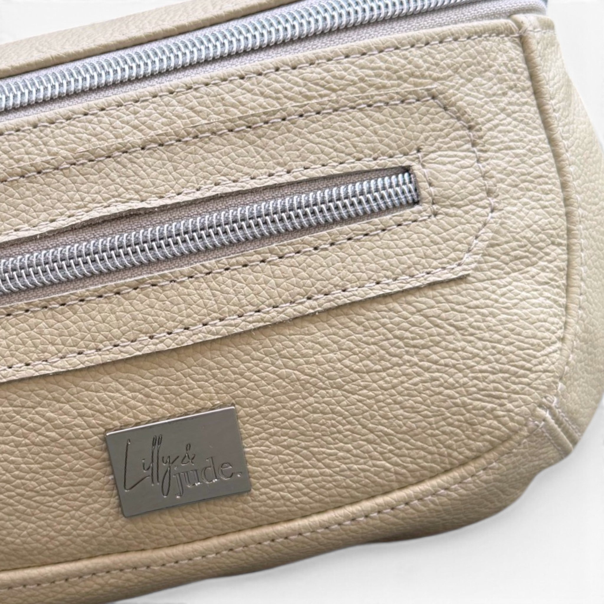 Close-up of a cream coloured pebbled leather bag with silver zippers and a silver metal brand plaque reading 'Lally Jude'.