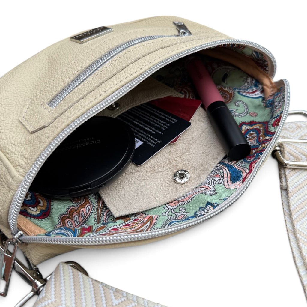 A close-up of a cream leather crossbody bag shown open, revealing a colorful paisley-patterned interior lining. Inside, a black compact, a card, and a pink lip product are visible.