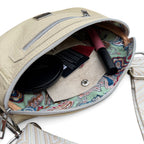 A close-up of a cream leather crossbody bag shown open, revealing a colorful paisley-patterned interior lining. Inside, a black compact, a card, and a pink lip product are visible.