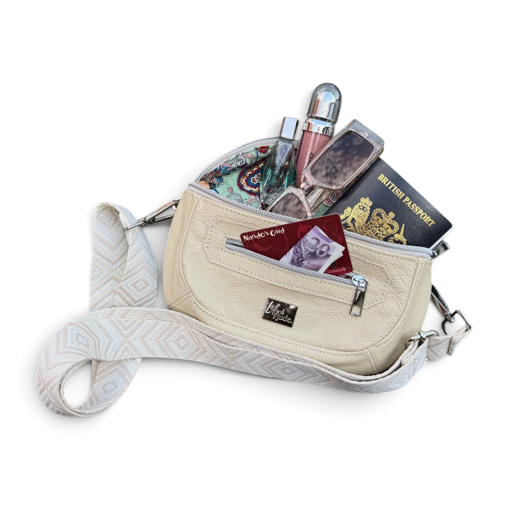 A cream-colored crossbody bag with a patterned strap, shown open and filled with various travel items including a lipstick, perfume, a passport, and a wallet, on a white background.