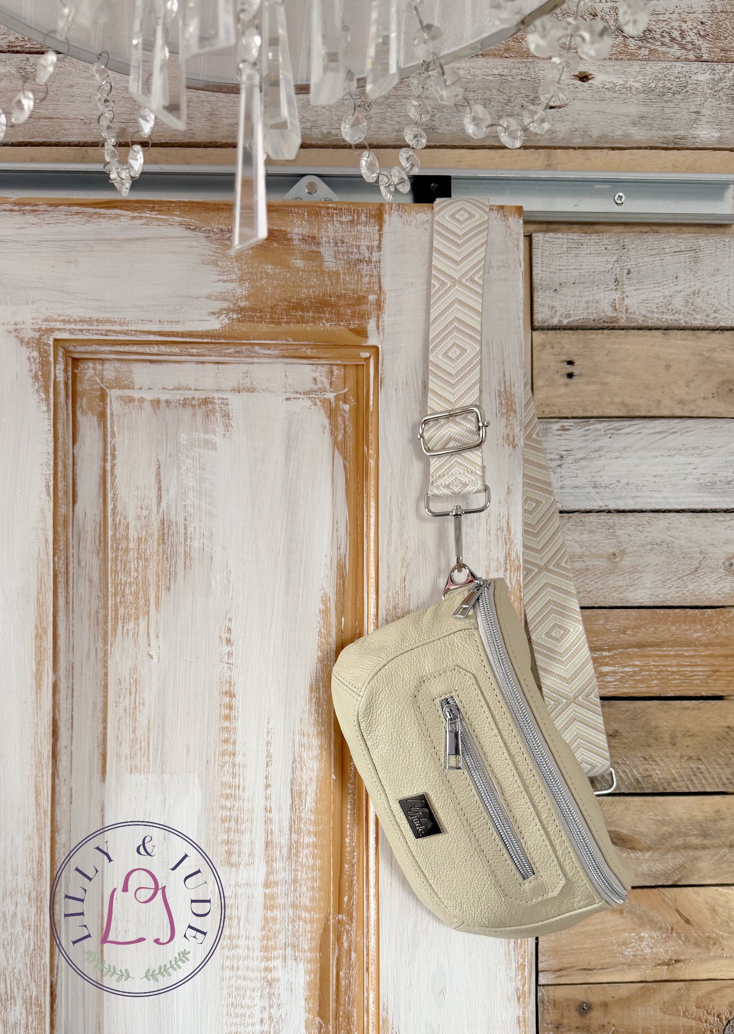 A cream-colored crossbody bag with silver zippers and a patterned strap, hanging on a rustic wooden door.