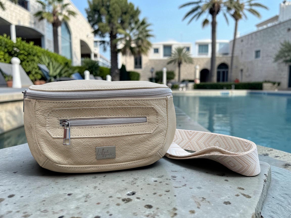 A cream coloured leather waist bag with a matching patterned shoulder strap, resting on a stone ledge in front of a resort pool with palm trees and buildings in the background.