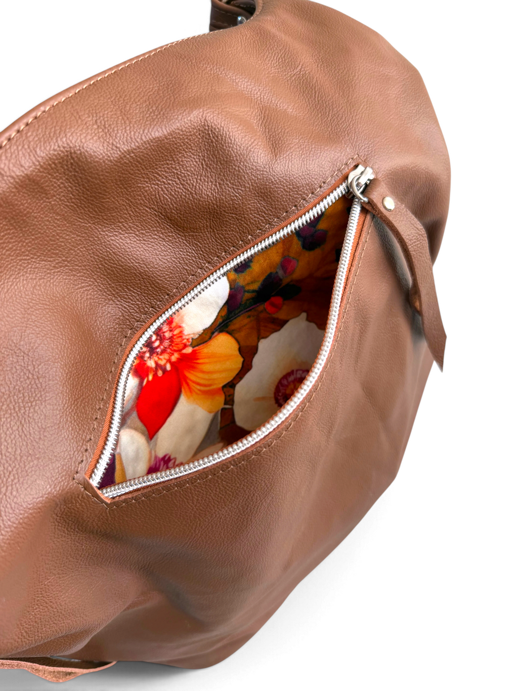 A close-up of a brown leather shoulder bag with an open zipper revealing a colourful floral-patterned interior, set against a plain white background.