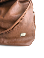 Close-up of a brown textured leather bag with a small silver-tone metal 'Lilly & Jude' logo plaque on the front, photographed against a white background.