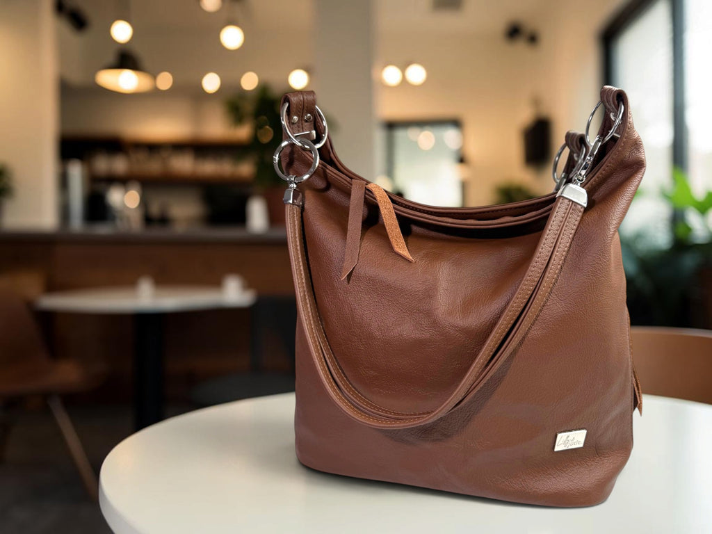A brown leather hobo shoulder bag with silver-tone hardware, displayed on a white table in a blurred cafe setting.