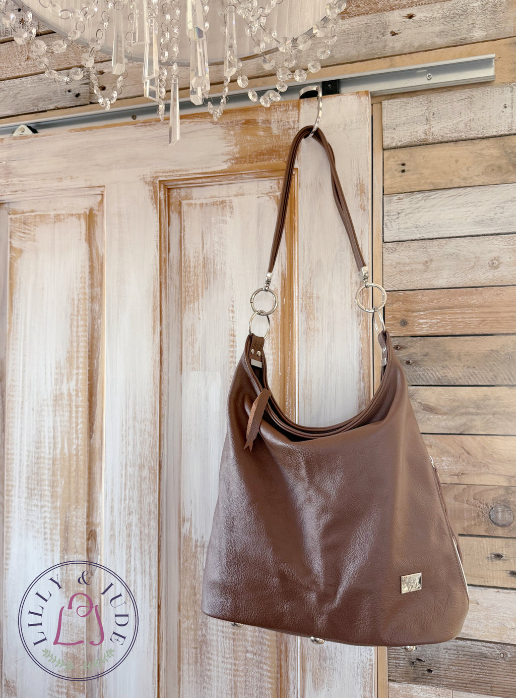 A brown leather slouchy shoulder bag with a strap and silver-tone hardware, hanging from a hook on a rustic wooden door with a chandelier visible above.