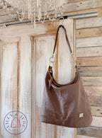A brown leather slouchy shoulder bag with a strap and silver-tone hardware, hanging from a hook on a rustic wooden door with a chandelier visible above.