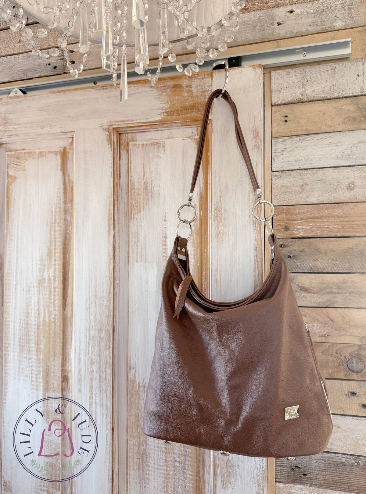 A brown leather slouchy shoulder bag with a strap and silver-tone hardware, hanging from a hook on a rustic wooden door with a chandelier visible above.