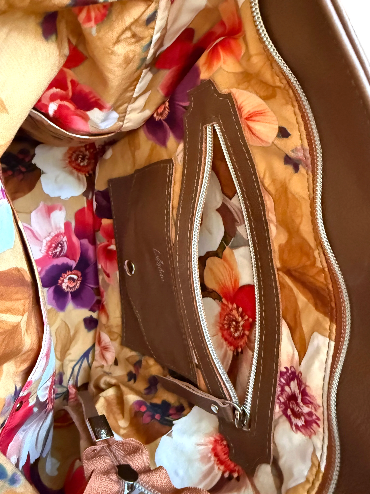 Close-up of the interior of a brown lrather tote, showing a floral-lined compartment with a zip pocket and a matching brown card holder. The lining has a warm tan base with large floral print in red, orange, purple, and white, while the bag's exterior trim is brown.