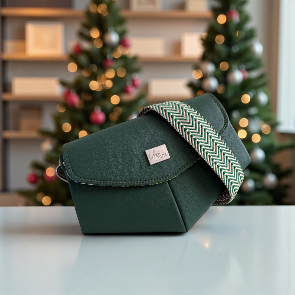 Green leather handbag with a chevron patterned crossbody strap on a table in front of decorated Christmas trees. The bag is a one-off edition and is made from sustainable materials