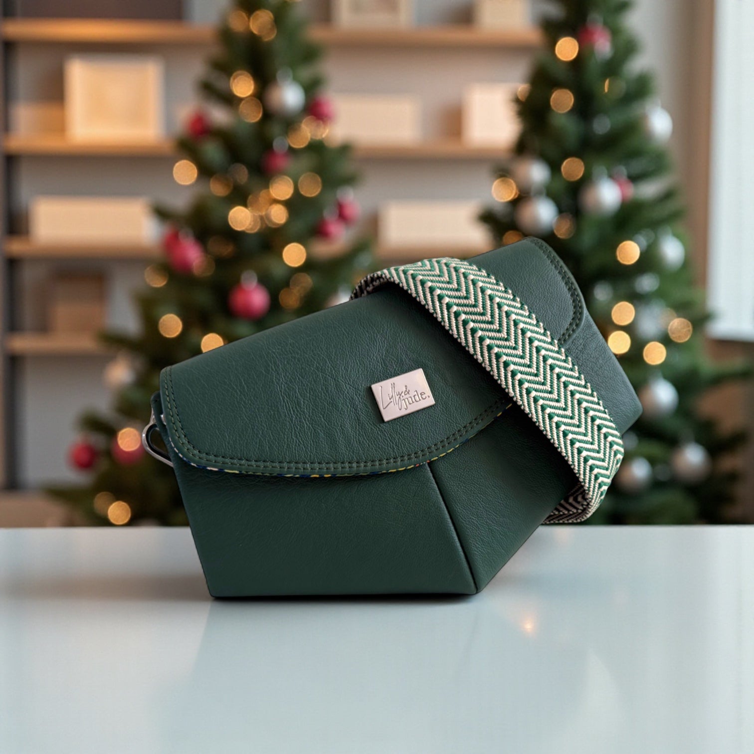 Green leather handbag with a chevron patterned crossbody strap on a table in front of decorated Christmas trees. The bag is a one-off edition and is made from sustainable materials