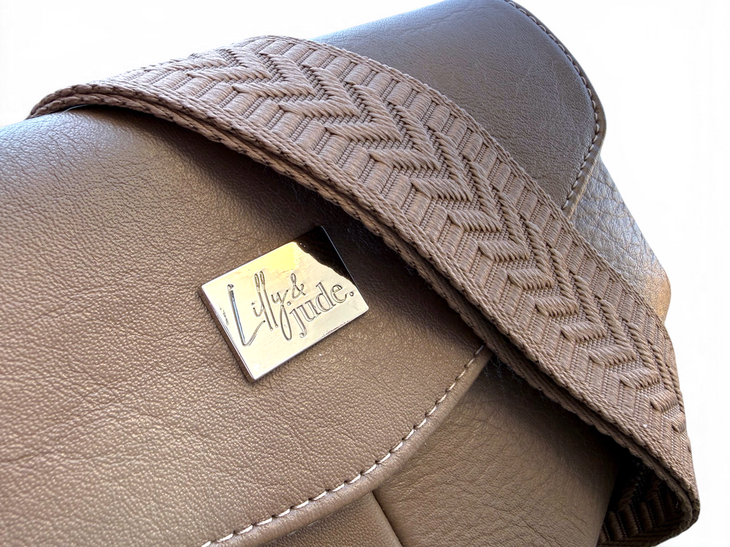 Close-up of a taupe leather bag with a woven herringbone strap and a silver-tone metal brand plaque reading 'Lilly & Jude'.