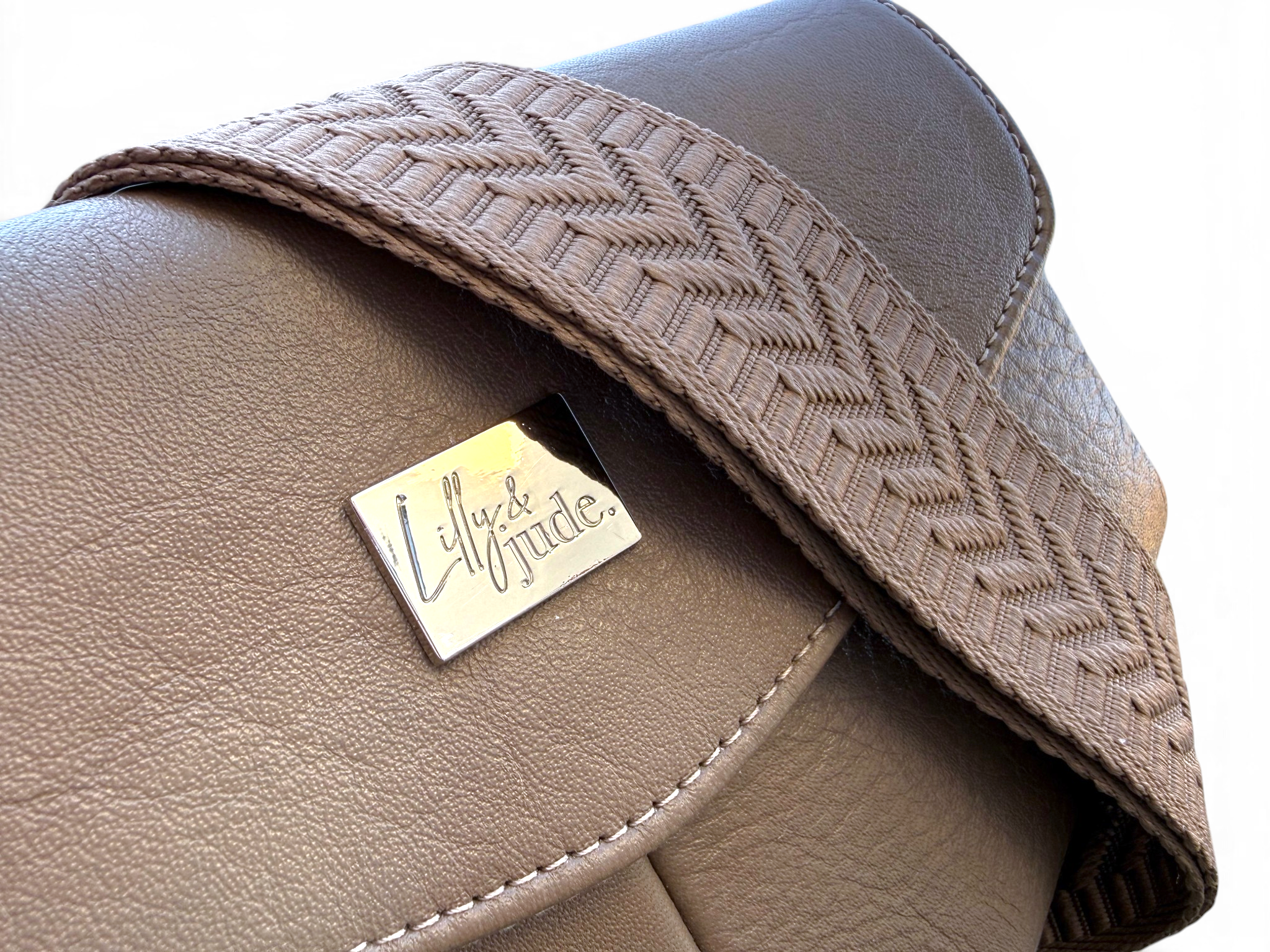 Close-up of a taupe leather bag with a woven herringbone strap and a silver-tone metal brand plaque reading 'Lilly & Jude'.