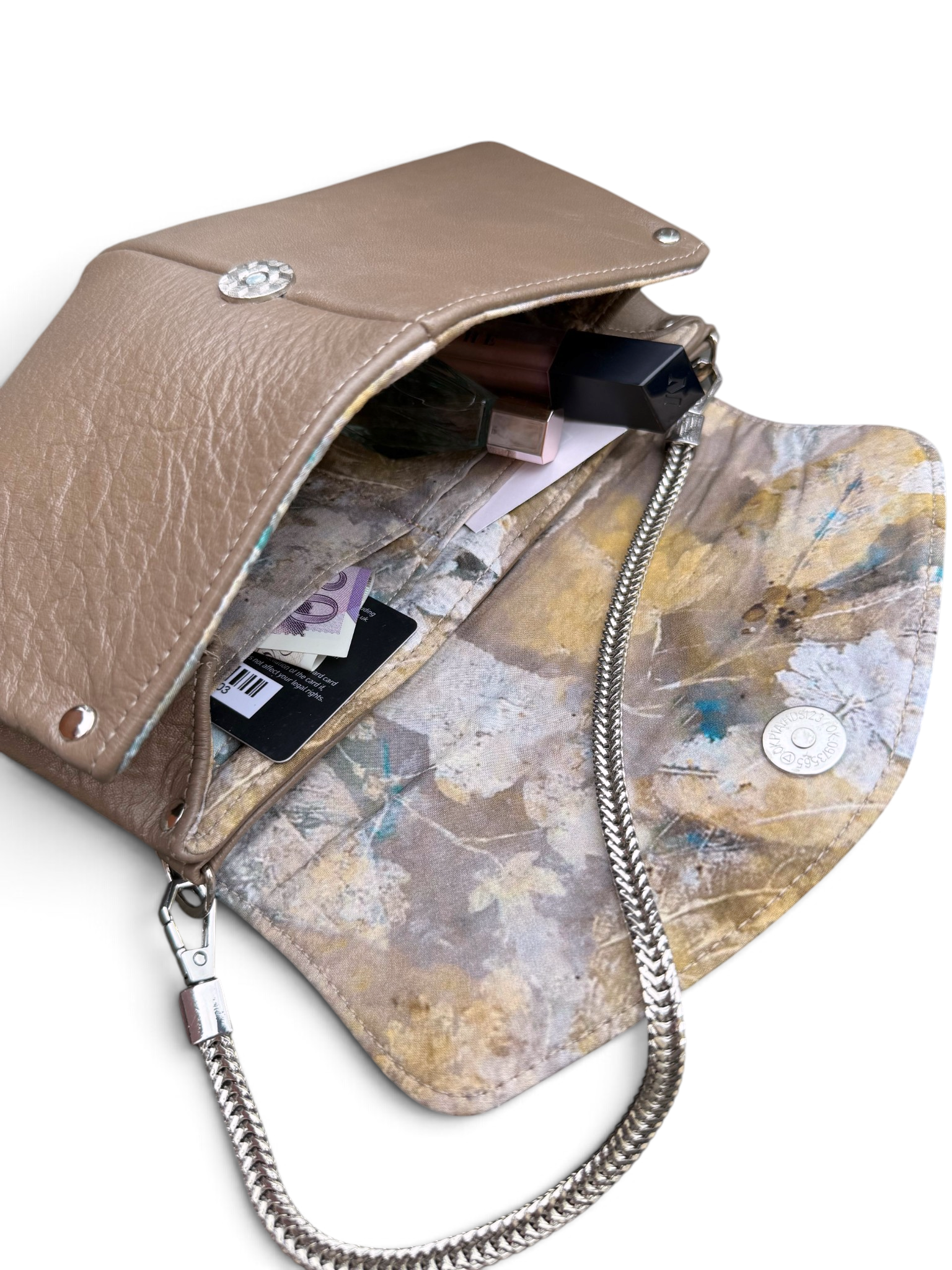 A tan leather clutch bag shown open, revealing a patterned interior with a silver chain strap, metal snap closure, and visible contents including cards and a lipstick