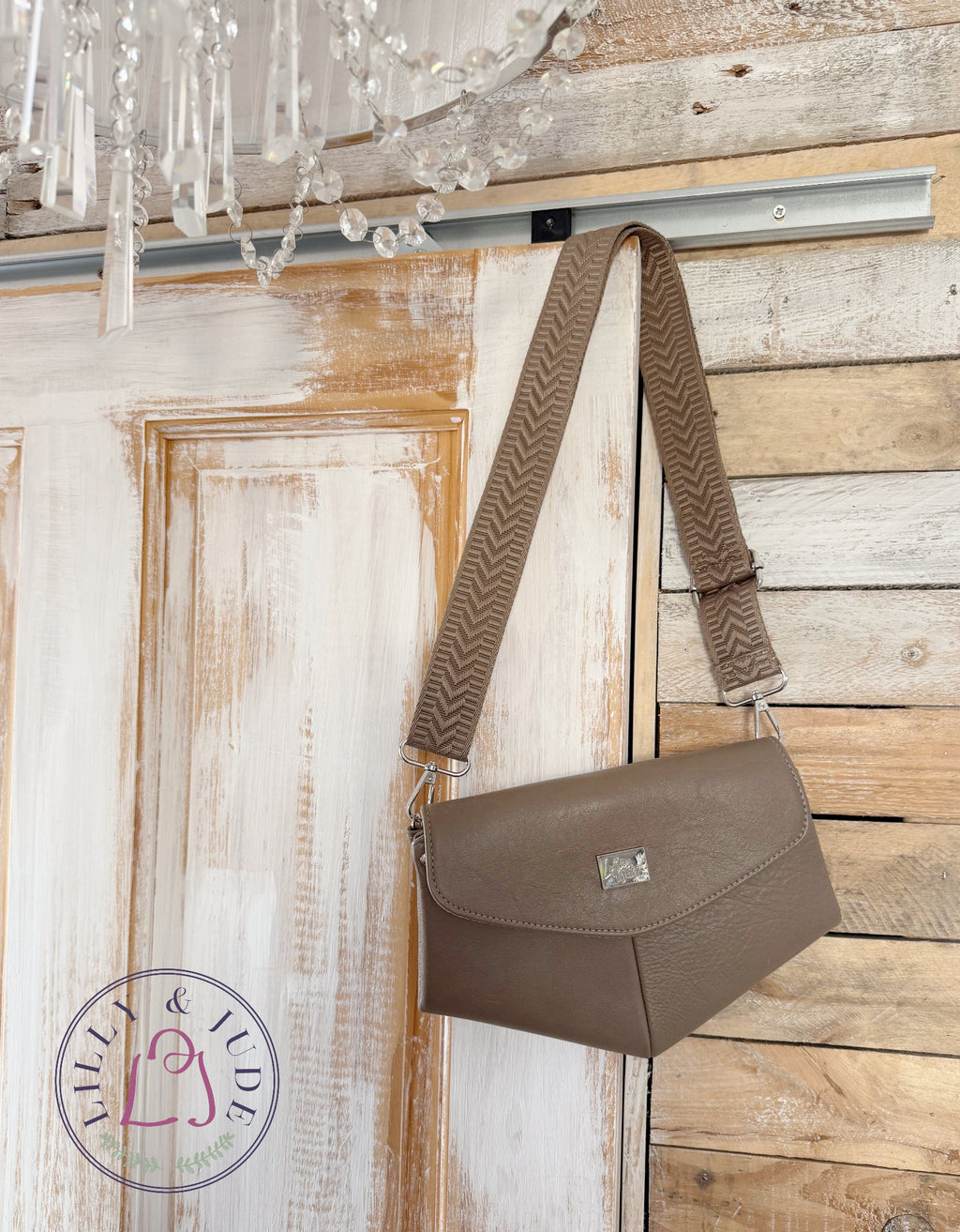 A taupe brown leather shoulder bag with a wide, patterned brown and beige herringbone strap, hanging from a silver rod against a rustic white and wood paneled wall.