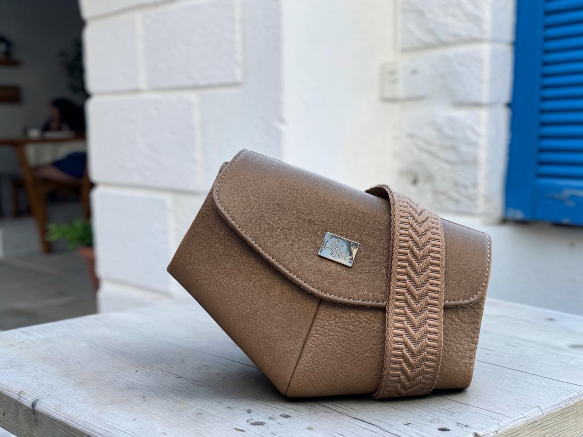 A taupe brown leather shoulder bag with a wide chevron-patterned strap, resting on a light wooden table in front of a white brick wall with a blue shutter.