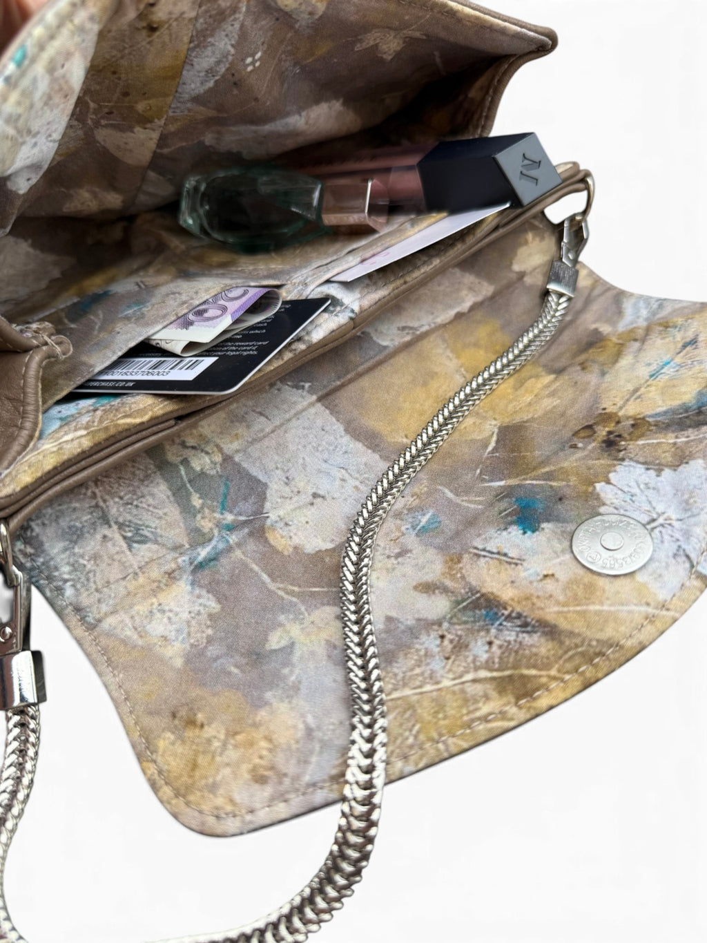 An open handbag with a distressed abstract patterned lining in beige, brown, and muted blue, featuring a silver chain strap and interior pockets containing cosmetics.