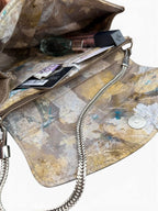 An open handbag with a distressed abstract patterned lining in beige, brown, and muted blue, featuring a silver chain strap and interior pockets containing cosmetics.