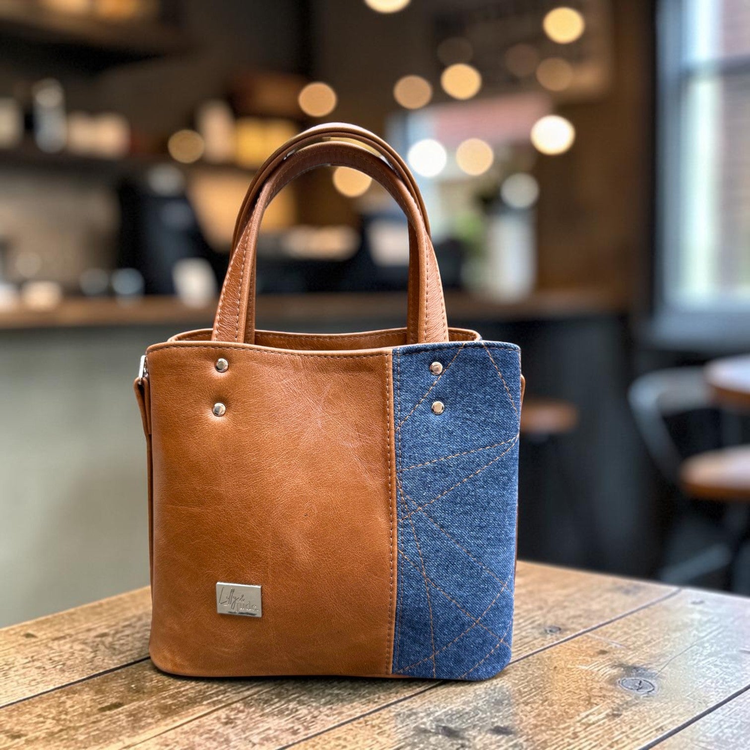 Two-toned tan brown leather and blue denim handbag on a wooden table with a blurred coffee shop background