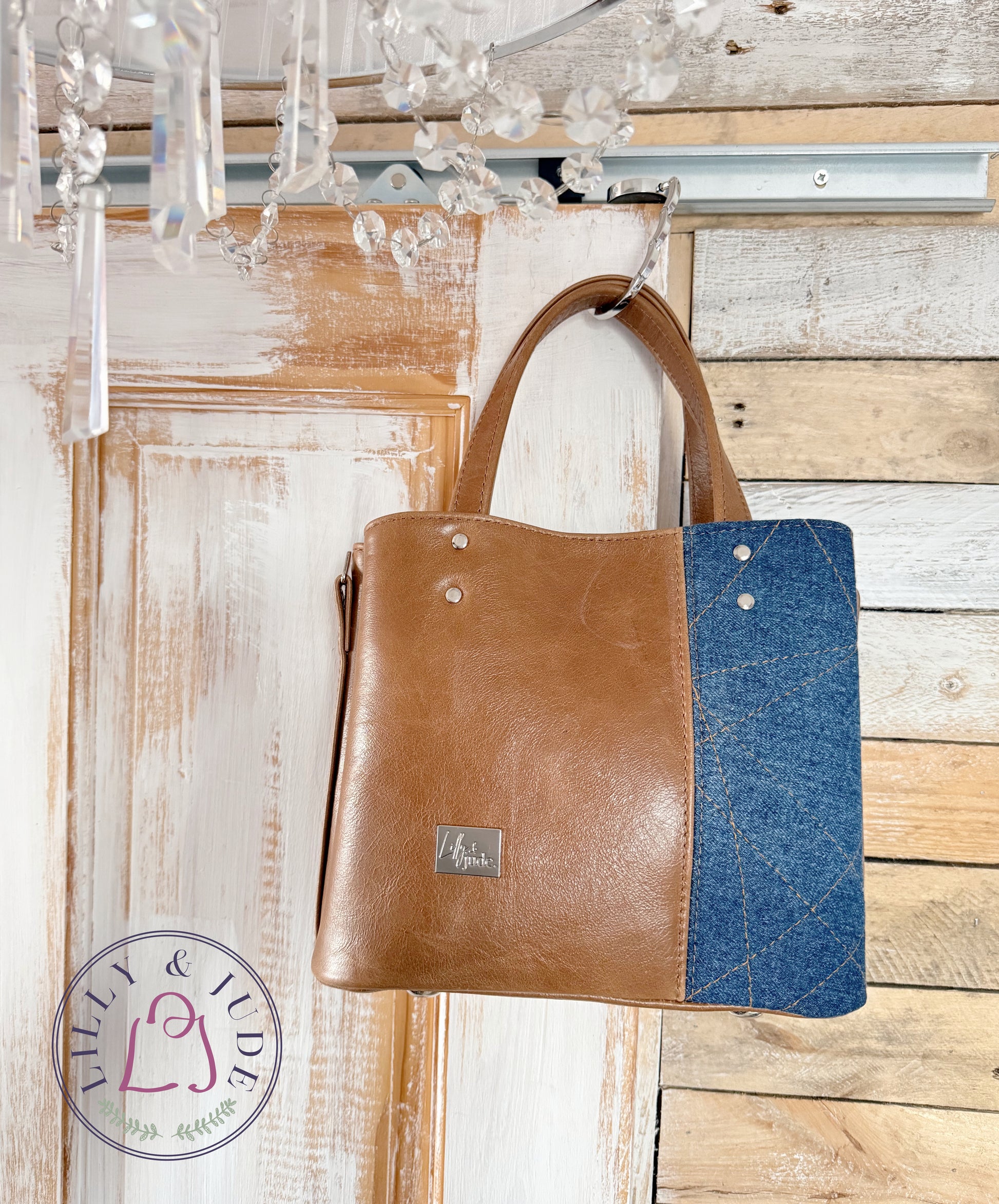 Brown leather handbag with blue denim section hanging on a wooden door.