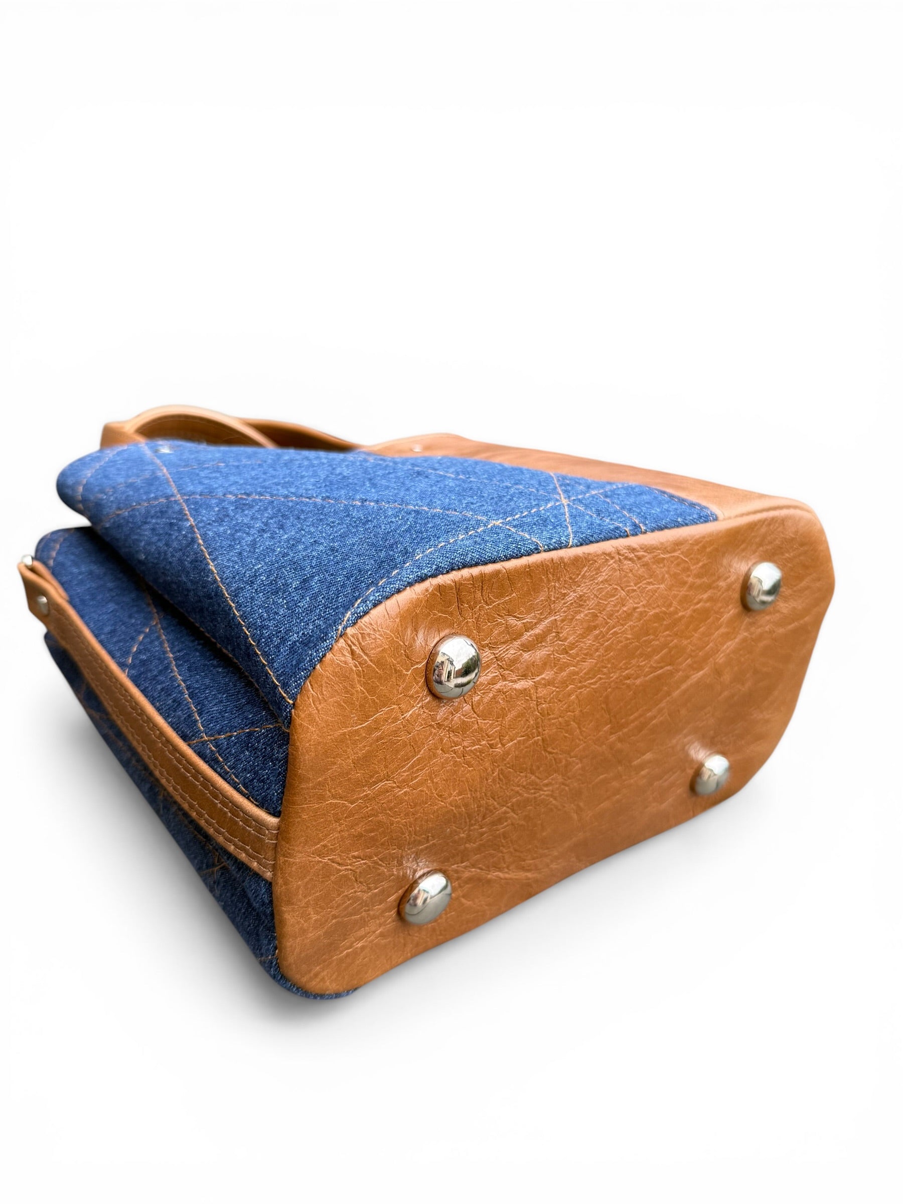 View of the base of the Soraya handbag, showing the tan brown leather base and 4 silver bag feet, and the denim panel on the bag, on a white background