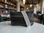 Black leather crossbody handbag with a decorative woven strap on a marble surface in a restaurant setting. Exclusive to Lilly & Jude