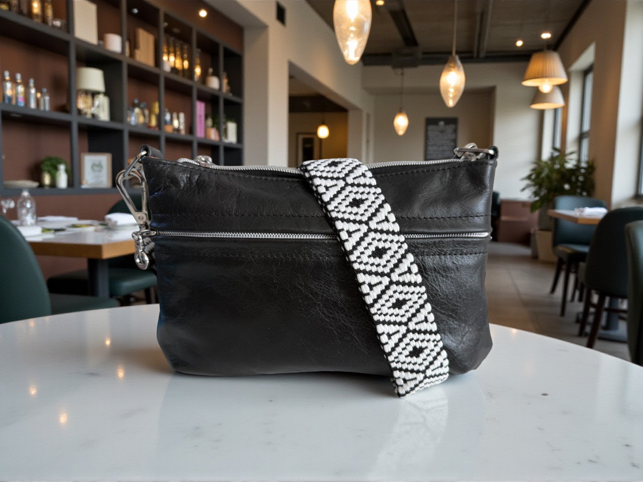 Black leather crossbody handbag with a decorative woven strap on a marble surface in a restaurant setting. Exclusive to Lilly & Jude