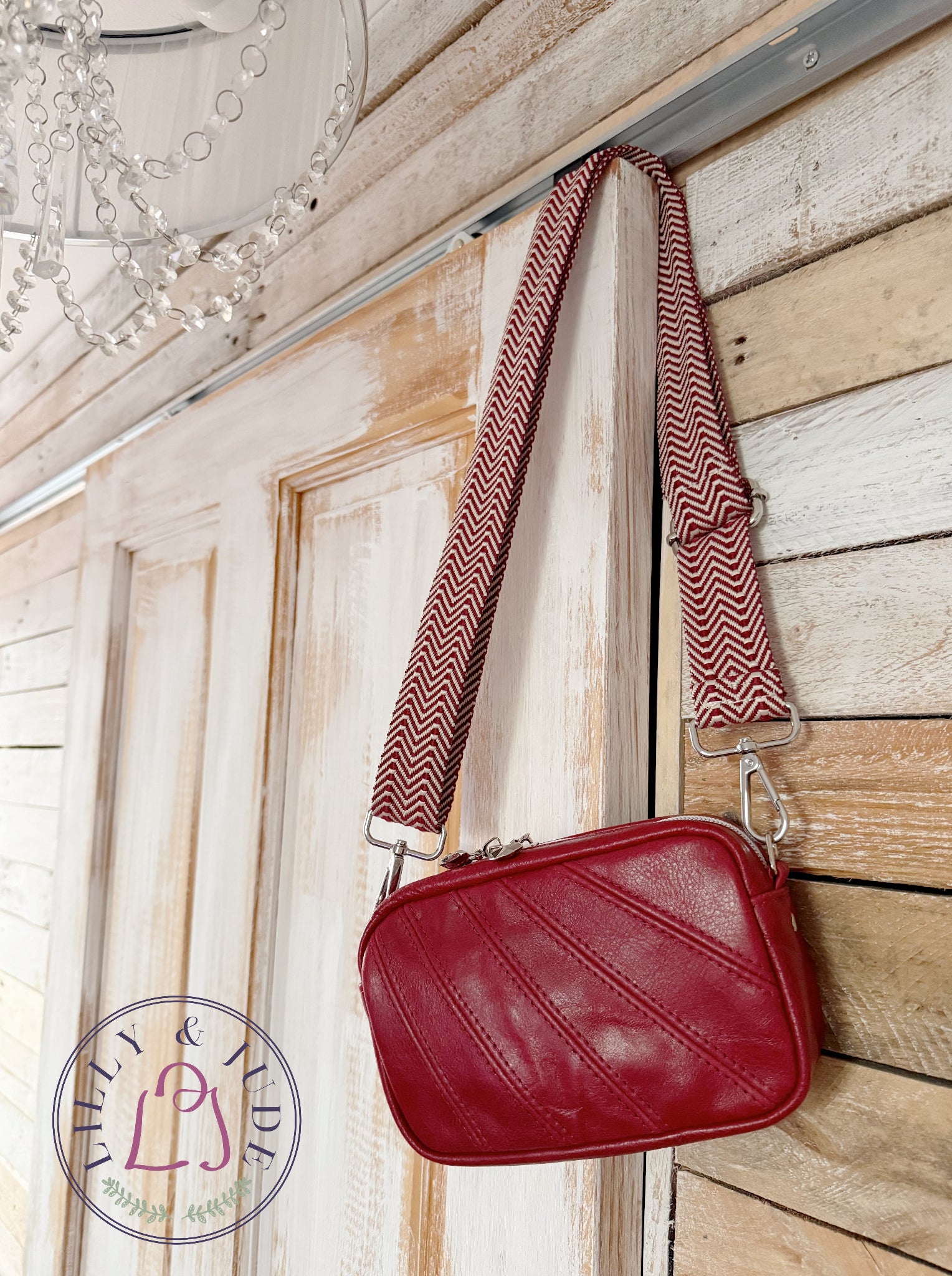 Red leather, compact crossbody bag, hanging from the top corner of a door, by its woven, chevron patterned strap. The image also has a Lilly & Jude logo in the bottom left hand corner