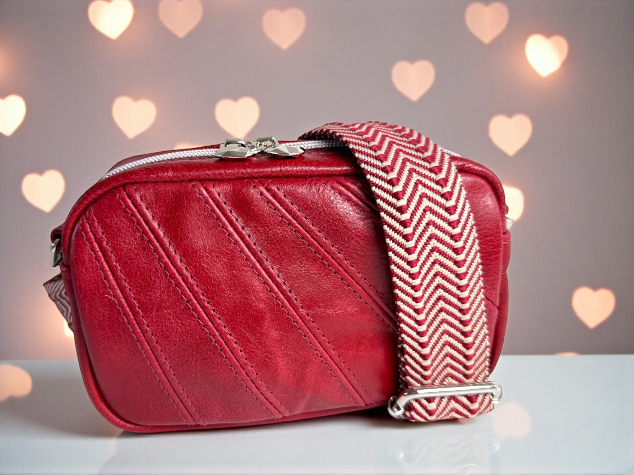 Red leather handbag with a chevron patterned strap on a light surface with heart-shaped lights in the background.