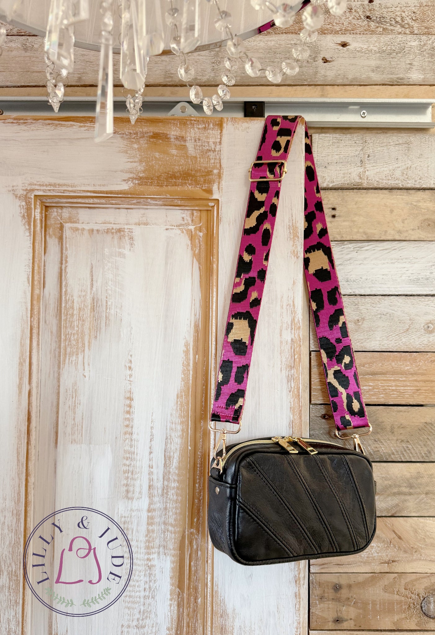 Small, black leather crossbody bag with a pink leopard print strap hanging on a wooden door.