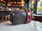 Compact black leather crossbody bag with a fuchsia leopard print strap on a table in a cafe setting