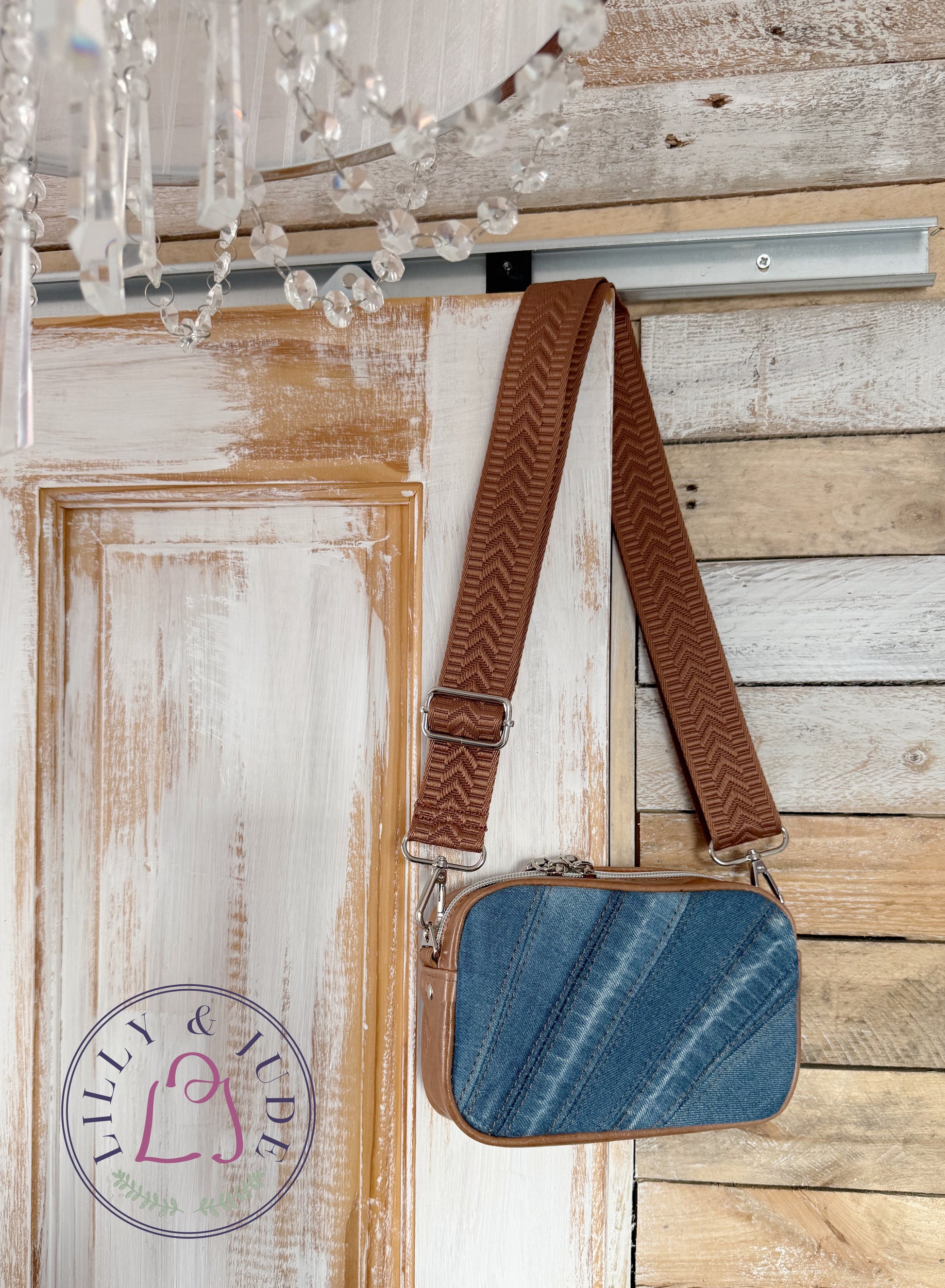 Blue upcycled denim bag with tan gusset and matching brown strap hanging on a wooden door, with Lilly & Jude logo