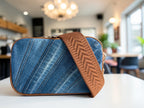 Blue denim crossbody bag with tan brown strap on a table in a blurred coffee shop setting