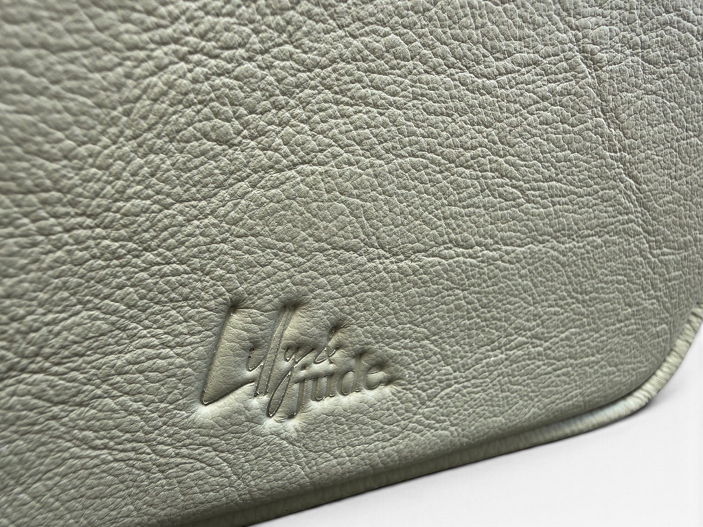 Close-up of a textured leather compact crossbody bag in a muted sage green color, featuring an embossed logo that reads 'Lilly & Jude'.