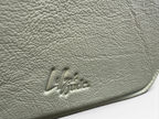 Close-up of a textured leather compact crossbody bag in a muted sage green color, featuring an embossed logo that reads 'Lilly & Jude'.