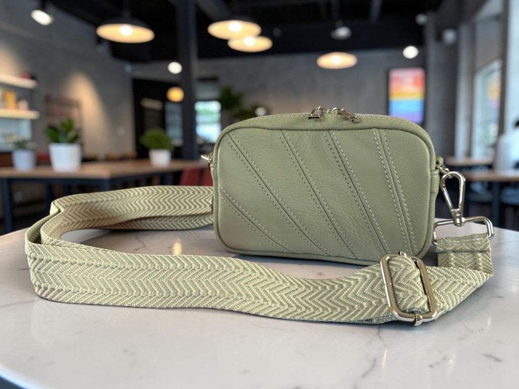 A sage green leather camera bag with diagonal stitched detailing, resting on a white marble countertop with its matching patterned shoulder strap spread out. The background shows a blurred cafe interior with tables and hanging lights.