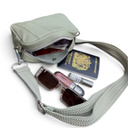 A sage green leather crossbody bag with a matching herringbone-patterned strap, shown open with its contents styled out: a UK passport, sunglasses, a lip gloss tube, and a small bottle. The background is solid white.