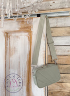 A sage green quilted crossbody bag with a matching woven shoulder strap, hanging on a rustic wooden door and wall. The bag features silver-tone hardware and a zip-top closure.