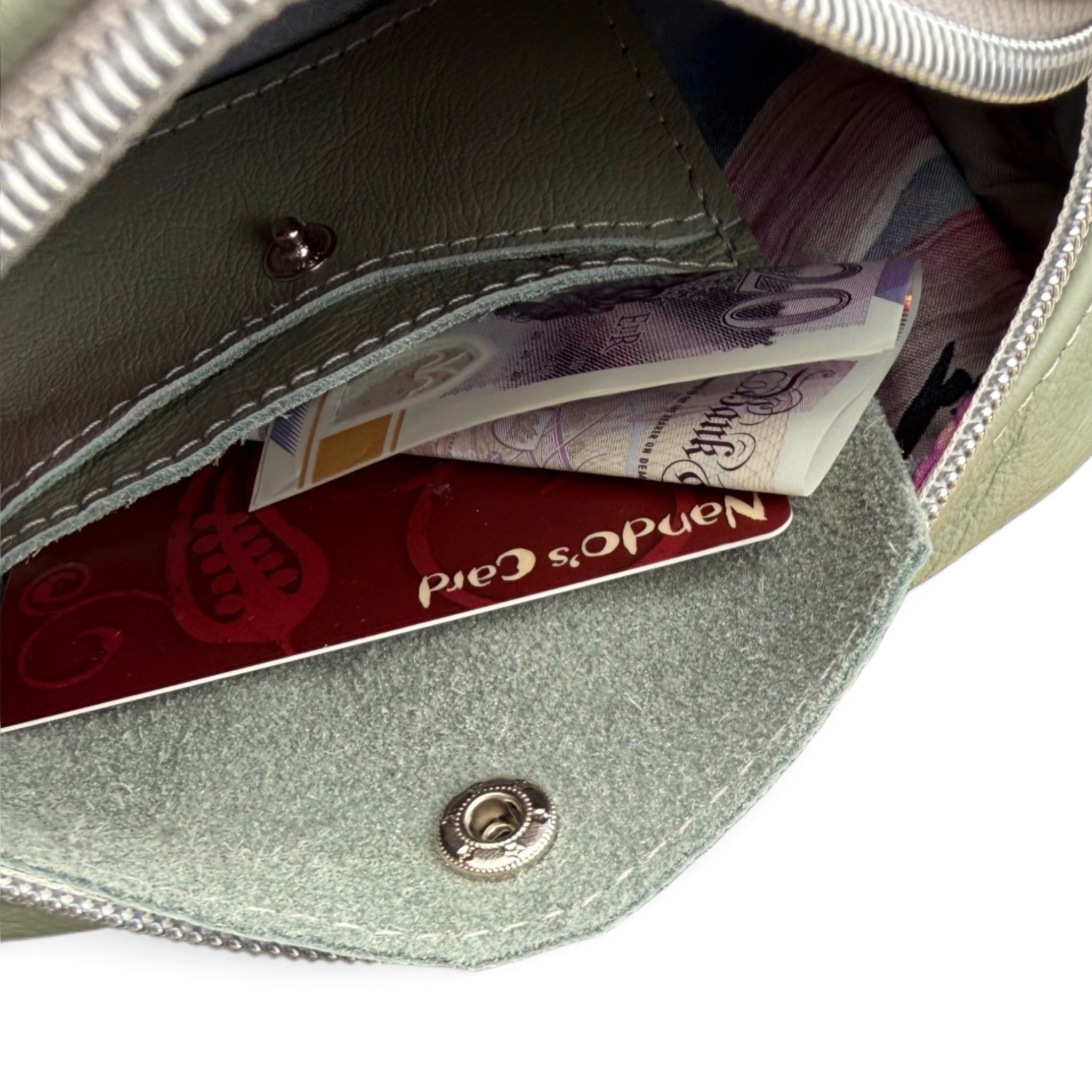 Close-up of the inside of a sage green leather zippered pouch, showing a silver zipper, snap button, card slots with a banknote and credit card, and a coin pocket on a white background.