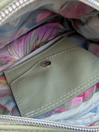 Close-up of the interior of a handbag or pouch, showing a light sage green leather integrated purse with white stitching and a metal snap, set against a pastel pink and blue floral fabric lining and a silver zipper.